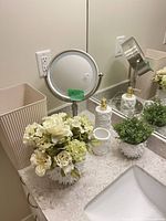 Countertop view showing lighted magnifying mirror, wastebasket, soap dispenser, tumbler, faux floral arrangements