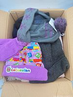 Open box showing multiple hats including grey pom-pom, purple Cocomelon beanie, dark knit items