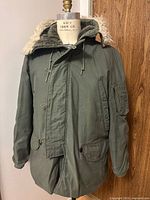 Front view of olive parka on mannequin showing hood, zipper, storm flap and pockets
