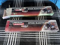 Two Lincoln Electric stick electrode boxes on blanket, one opened, one sealed