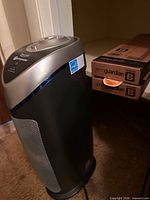 Tower air purifier standing on floor next to two boxed replacement filters on shelf