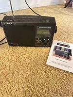 Front view of C Crane CC Radio 2E showing speaker, display, controls and antenna with manual beside