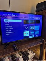 Front view of Insignia 32" Roku TV on stand displaying home screen; remote visible on shelf