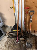 Group of long-handled tools leaning against wall: leaf rake, rotary cultivator, hoe, cultivator fork, two shovels