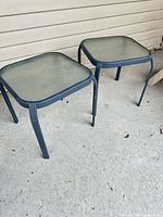 Two square outdoor patio tables with glass tops and metal legs