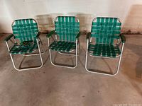 Three green webbed aluminum lawn chairs aligned side by side on concrete floor