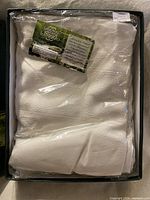 Folded white/off-white linen tablecloth in plastic sleeve inside box with hang tag visible