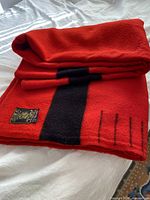 Blanket folded on bed showing red wool, central black stripe, stitched point marks and label