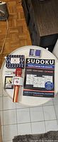 Top-view of all five items on round table: Sudoku book, wireless karaoke microphone, playing card deck, small cassette-style card pack, and compact boxed game