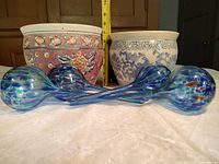 Two ceramic planters side by side with four blue/teal speckled glass watering bulbs laid in front, tape for scale