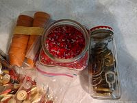 Group shot of square glass jar with red beads, tall glass jar with metal hardware, stack of terracotta pots bound by elastic, partial view of zipper bags with buttons