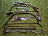 All tools laid out on green surface showing hand saws, bow saws, hacksaw, hatchet, axe head, loose blades and wedges