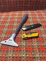 Husky wall scraper, Husky black utility knife, DeWalt yellow utility knife on red-brown patterned surface