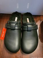 Top view of black clogs showing uppers, buckle, faux-fur lining, hang tag