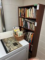 Bookcase filled with assorted books, wicker basket on top of dryer, large art book in foreground