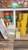 Cabinet shelf showing yellow canisters, mixing bowls, cups, cereal dispensers, pitcher and floral tray