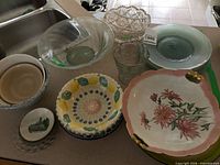 Group shot on countertop showing glass bowls, green plates, souvenir plate, speckled plate, beige bowl, hand-painted bowl, pink floral plate