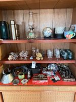 Overall lot on shelving showing thermos, bottle, oil lamp, globe, glassware, ornaments, cookware