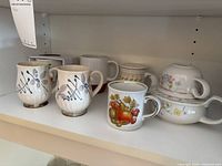 Shelf view showing five distinct mugs, fruit motif mug, two blue-pattern mugs, plain mug, floral band mug, and teapot set