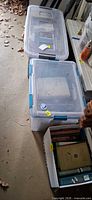 Two clear plastic lidded totes and one cardboard box filled with hardback books
