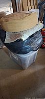 Clear plastic tote with black bag holding rugs, folded rag rug, and round burlap cushion stacked on top