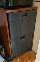 Cabinet front showing wood frame, two grey perforated metal drawers, mounted on casters