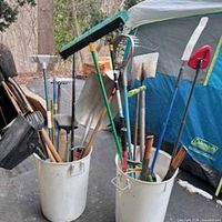 Two white buckets holding assorted long-handled yard tools such as shovels, brooms and snow shovel
