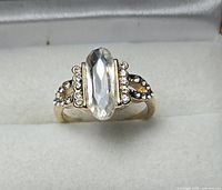 Gold-tone ring with elongated clear faceted center stone and pavé accents displayed on ring pad