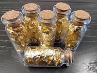 Front view of six glass vials filled with gold leaf flakes, one vial lying to show contents