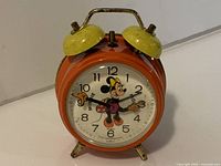 Front view of orange twin-bell alarm clock showing Minnie Mouse dial and brass feet