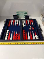 Open backgammon case showing board, checkers, dice, cups, instruction sheet, tape measure for scale