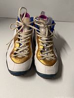 Front view of Nike Air Foamposite sneakers showing laces, gold and white uppers, blue trim