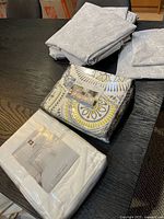 All bedding items together on table: two folded grey paisley sets, packaged white UGG set, packaged medallion queen duvet cover