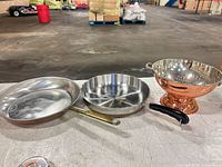 Three cookware pieces laid out: two frying pans and copper colander