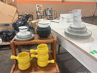 Overview of lot showing yellow handled bowls, black plates, patterned plates, white mugs and gold rim plates