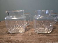 Front view of both clear pressed glass jugs showing square bodies and starburst pattern