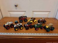 All nine monster trucks lined on wooden surface with ruler for scale