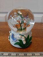 Front view of snow globe showing butterflies and lily-of-the-valley inside, sculpted base details visible