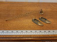Candle snuffer and two brass slippers on wood surface with ruler for scale
