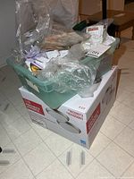 Green plastic tote of mixed hardware and plumbing accessories sitting on top of boxed LED ceiling fixture