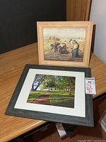 Two framed prints on desk surface: park pathway print in dark grey frame with white mat; harvest scene print in light wood frame