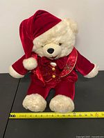 Front view of cream plush teddy bear in red velvet Santa outfit next to measuring tape
