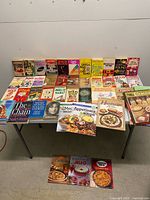 Wide view of entire lot arranged on table and floor showing quantity and variety of books and cookbooks.