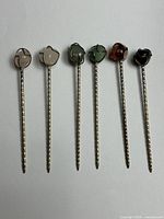 Six twisted metal cocktail picks with gemstone tops laid out side by side