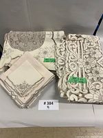 Folded linens: large embroidered tablecloth, smaller lace appliqué cover, stack of embroidered napkins