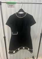 Front view of black mini dress on hanger showing gold trim, faux pocket and floral hem appliqués