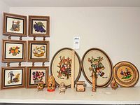 All items grouped on shelf: framed crewel embroideries, Bulgarian plate, Matryoshka dolls, wooden eggs, sphere, carved figurine