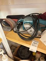 Makita belt sander with coiled cord, sanding belt and dust bag visible on shelf