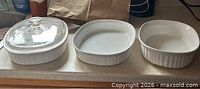 Three white CorningWare oval casserole dishes lined on countertop, leftmost with glass lid, center without lid, rightmost smaller dish