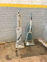 Both upright vacuum cleaners standing side by side against brick wall